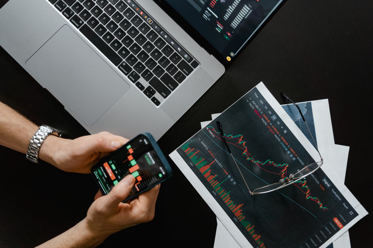 hero-img-01 Hands using smartphone beside laptop with stock charts, showcasing digital trading.